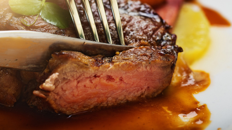 A knife and fork slicing into a filet mignon with sauce beneath it
