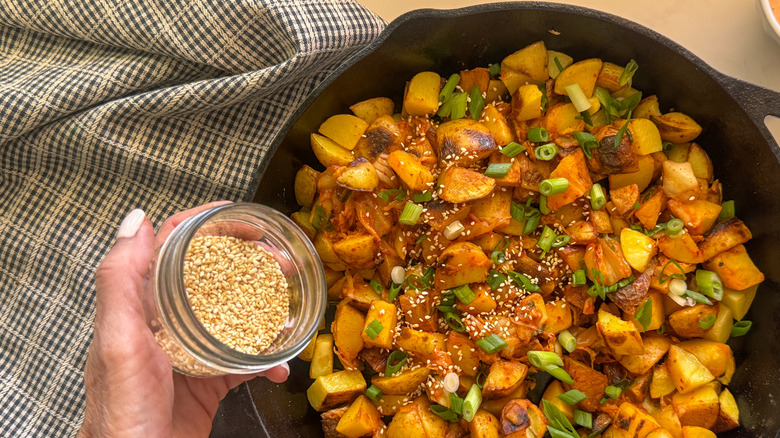 hand adding sesame seeds to potatoes in skillet
