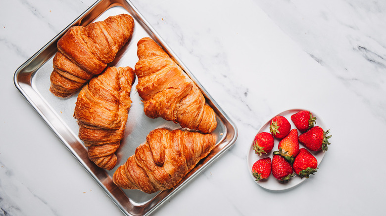 Four croissants on metal sheet besides strawberries.