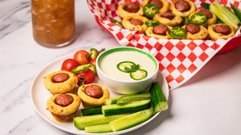 Mini corn dogs on plate with fresh cucumbers and jalapeño crema, with iced tea and more corn dogs in basket in background.