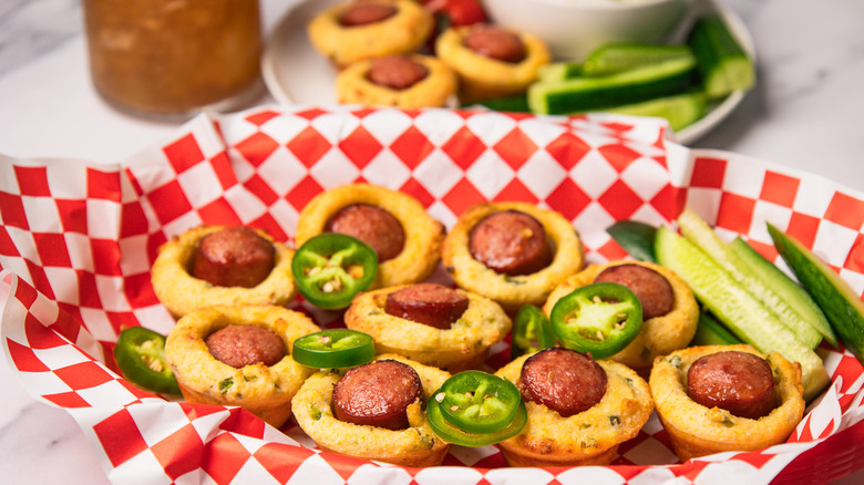 Mini corn dogs in basket with fresh cucumbers and jalapeños, and more on a plate in background.
