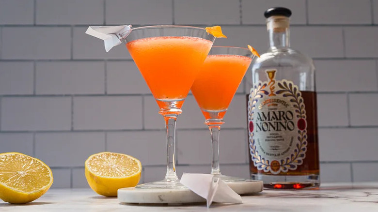 Two martini glasses with Paper Plane garnishes next to bottle of Amaro Nonino and halved oranges.