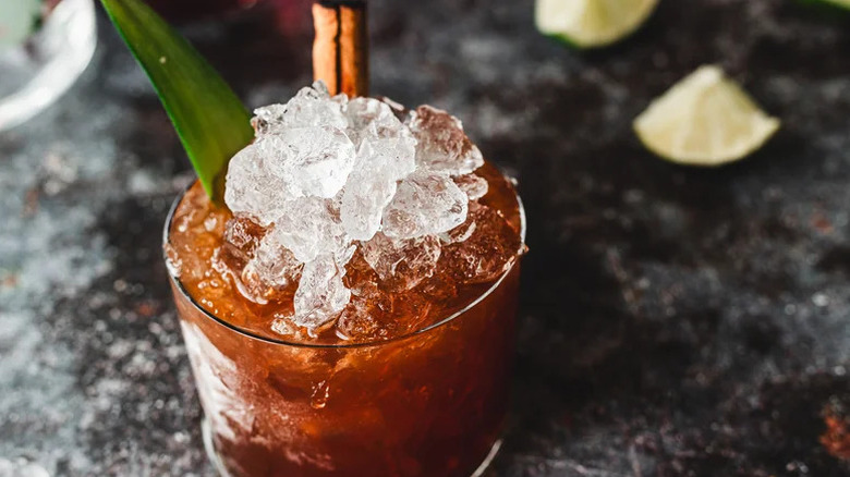 A close-up shot of a brown cocktail with crushed ice, cinnamon stick, and pineapple leaf garnish with lime wedge in background.