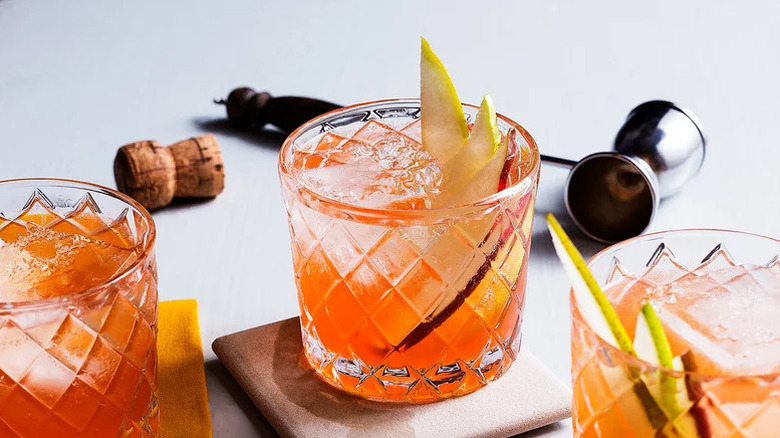 Three cocktails in old-fashioned glasses with sliced pear garnish, a jigger and cork against white background.