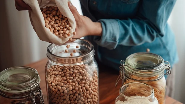 Closeup of woman pouring a bag of chickpeas bought from zero waste grocery store into glass jars. 