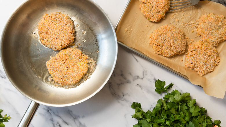 Crab cakes in a pan with more on baking sheet besides.