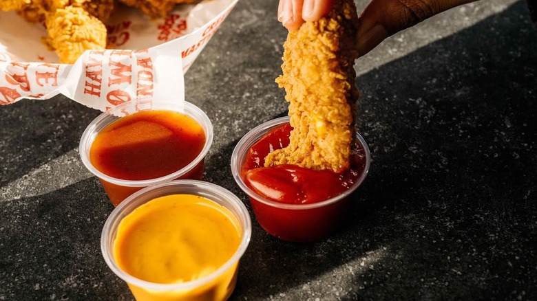Someone dipping a chicken tender into a small cup of ketchup, that's beside a basket of tenders and two sauce cups
