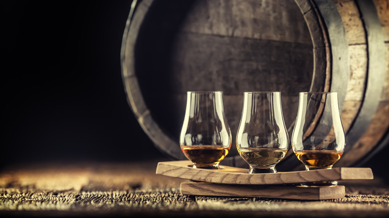 Three filled glasses of whisky on a plank with barrel and grains in the background