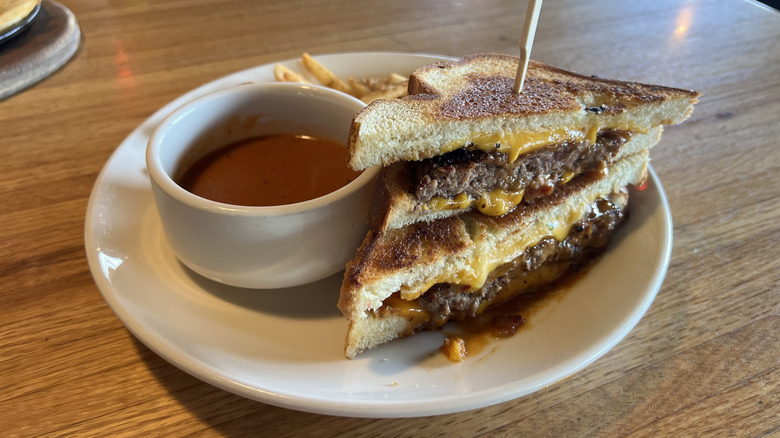 A white plate containing an Applebee's Grilled Cheese Cheeseburger and side of tomato soup and fries