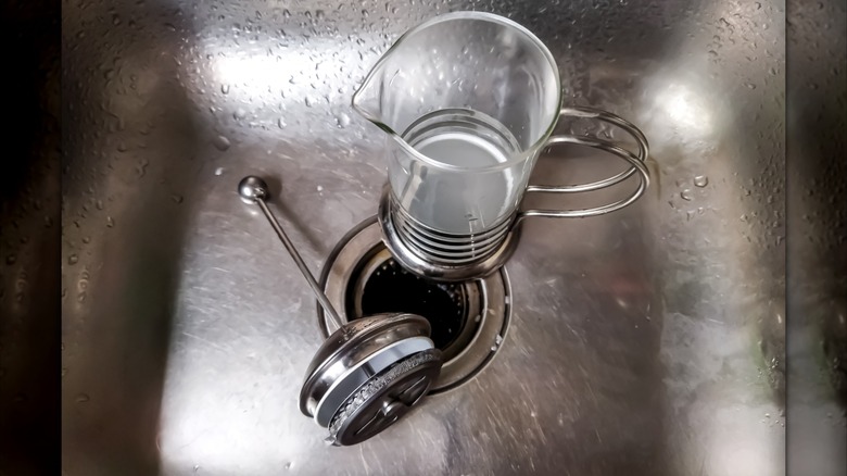 A French press jug and a plunger pictured in a stainless steel kitchen sink.