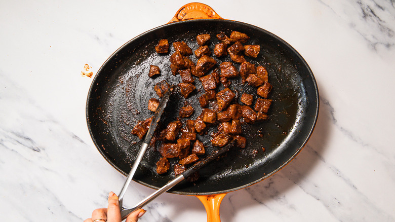 Tongs tossing steak bites in cast iron pan