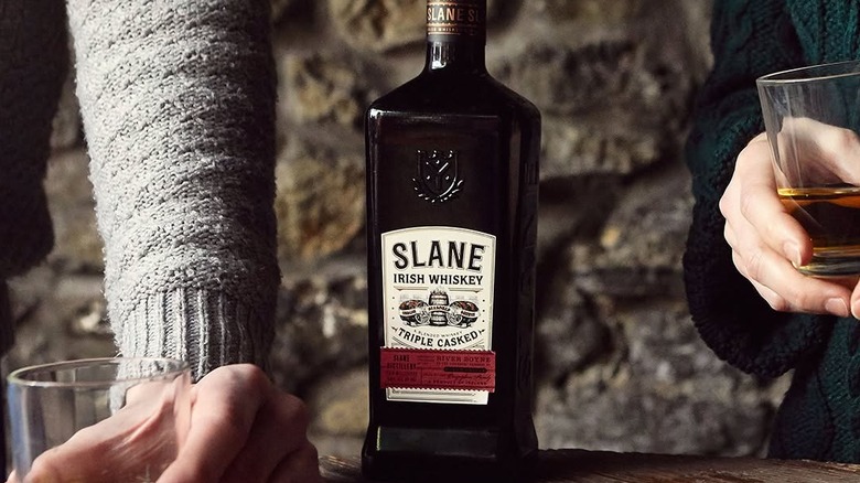A bottle of Slane Triple Casked Irish Whiskey on a table in front of a brick background, with people holding glasses on either side
