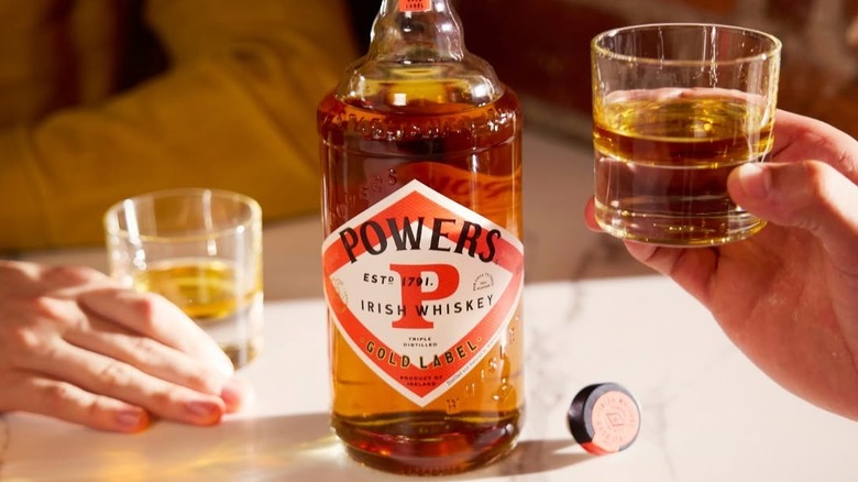 An open bottle of Powers Gold Label, with people holding filled glasses on either side