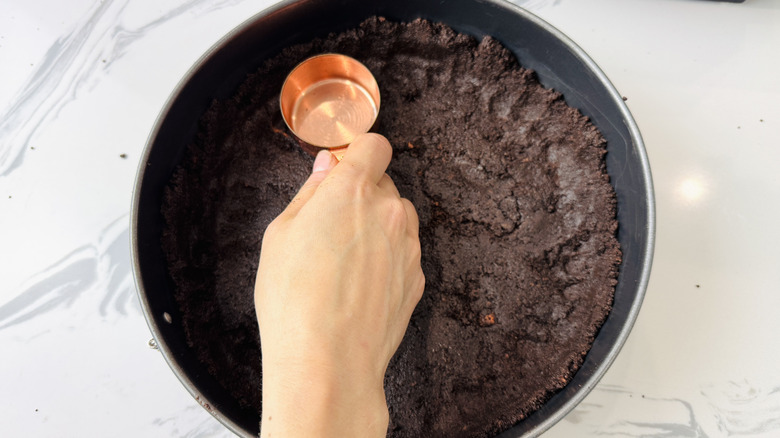 Hand using measuring cup to press Oreo crust into pan