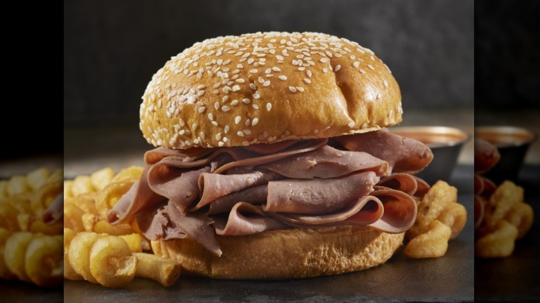a close-up shot of a tall stack of thinly sliced roast beef on a sesame seed bun with curly fries beside it