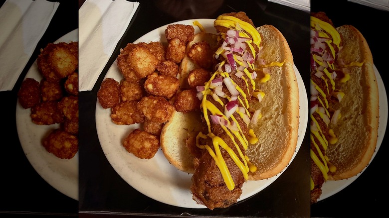 Deep-fried hot dog drizzled with sauces next to tater tots on white plate