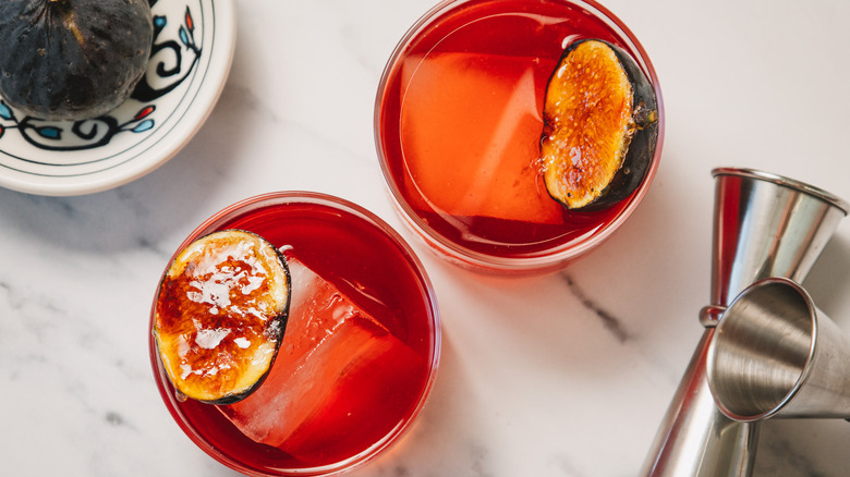 Overhead view of two cocktails with jiggers and whole fig
