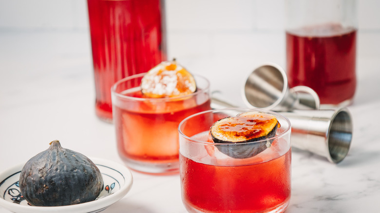 Two cocktails with jiggers and whole fig, bottles in background