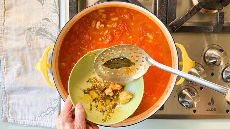 Using a slotted spoon to remove parmesan rind, thyme stems, and bay leaf from white bean tomato soup