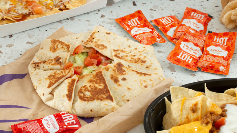 Taco Bell Crunchwrap Supreme resting on its paper bag surrounded by hot sauce packets and other menu items