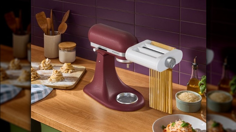 A dark red KitchenAid mixer with a pasta roller attachment on a wooden counter top