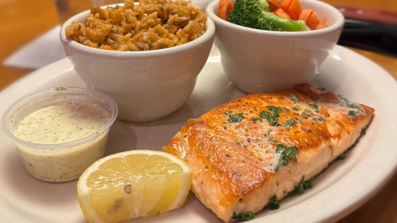 A plate of grilled salmon beside tartar sauce, a lemon wedge, and a bowl of veggies and a bowl of rice