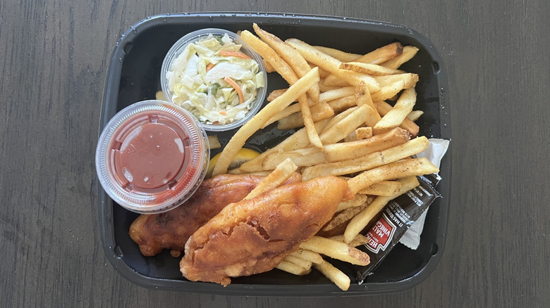 Black tray of Appleebee's fish and chips with fried fish, french fries, cup of cocktail sauce, and cup of coleslaw