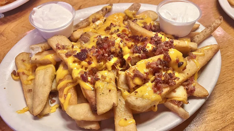 A plate of steak cut fries covered in melted cheese and bacon bits with two containers of sour cream